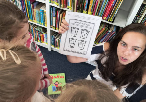 Pani bibliotekarka pokazuje kartę z koszami do segregacji śmieci.