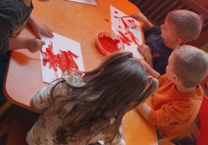 Grupa dzieci maluje czerwoną farbą kartki papieru.